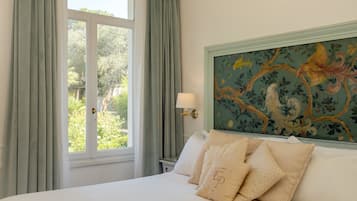 A bedroom with a large bed, a window with curtains, a nightstand with a lamp, and a wall with a floral painting.