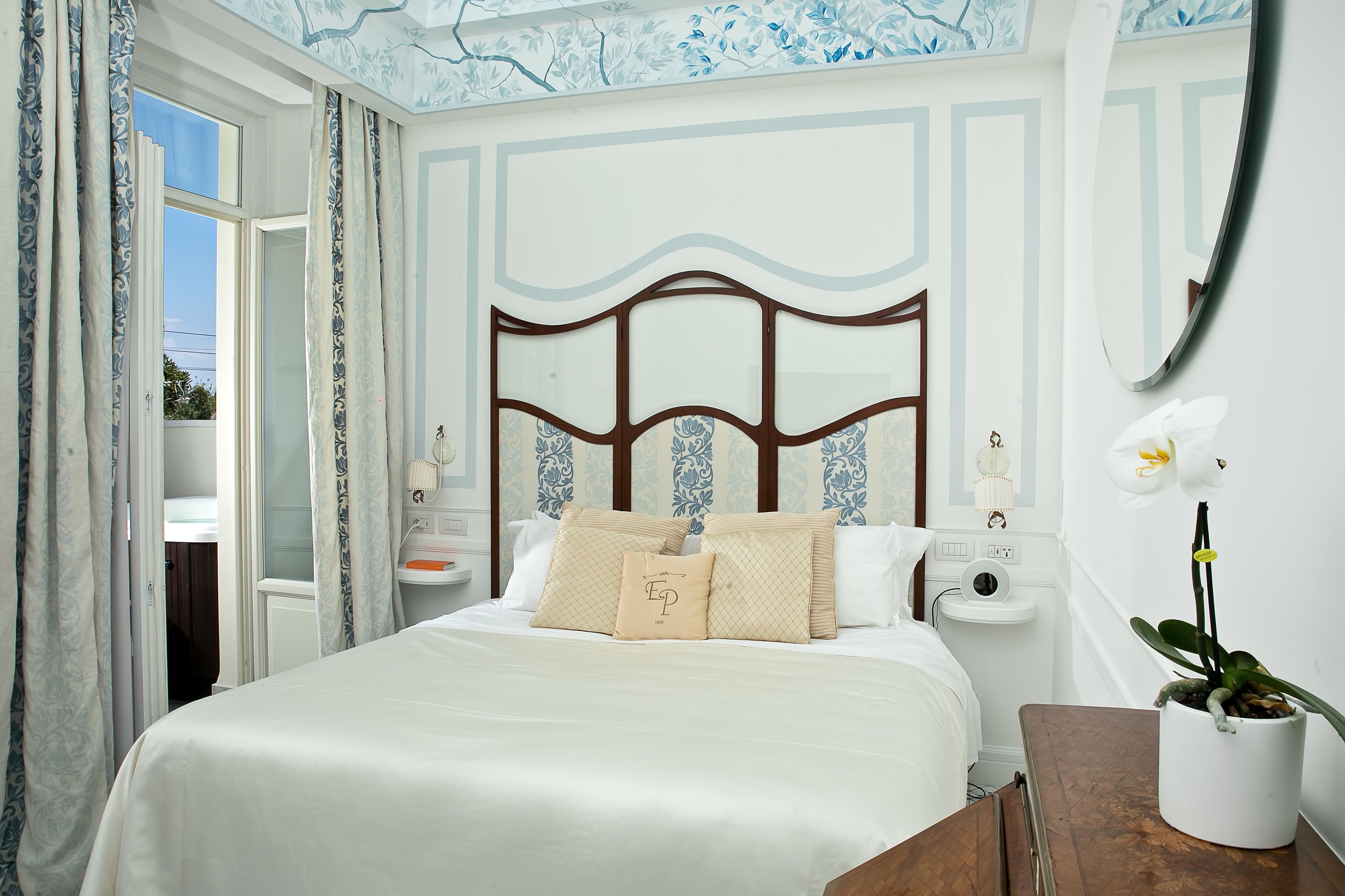 A bedroom with a large bed, a bedside table, a nightstand, a mirror, and a potted orchid.