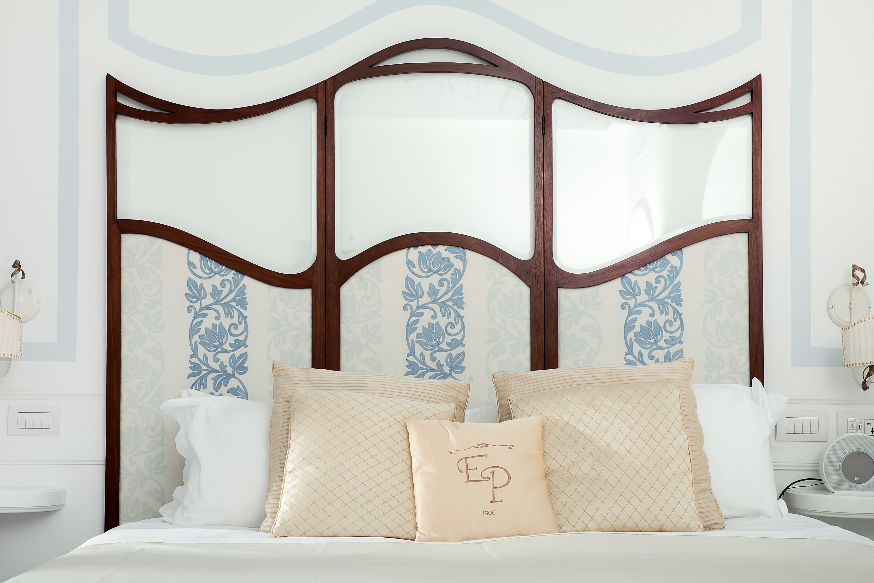 A bedroom with a large bed, a wooden headboard with glass panels, and patterned wallpaper.