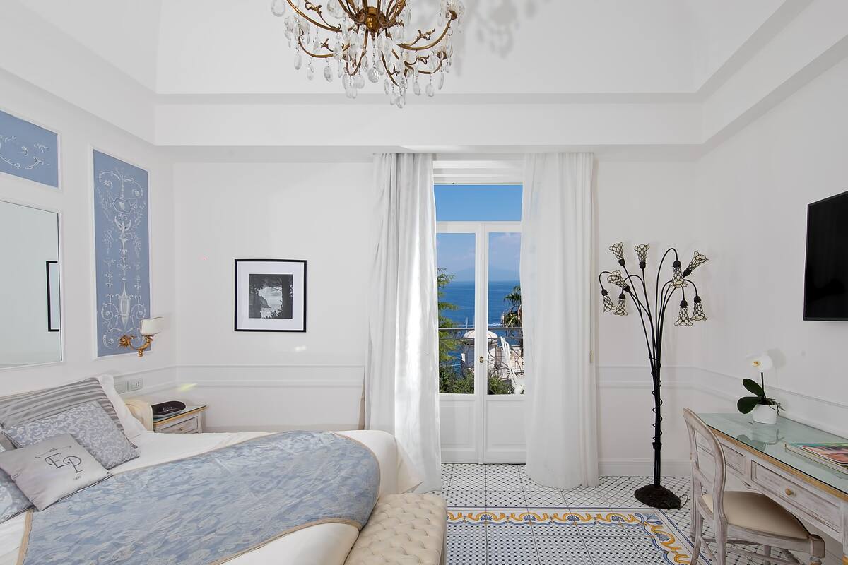 A bedroom with a bed, a desk, a chair, a chandelier, a floor lamp, and a view of the sea.