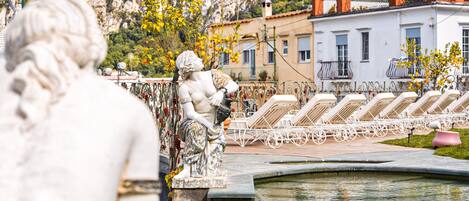 A statue in a pool area with lounge chairs and a building with balconies in the background.