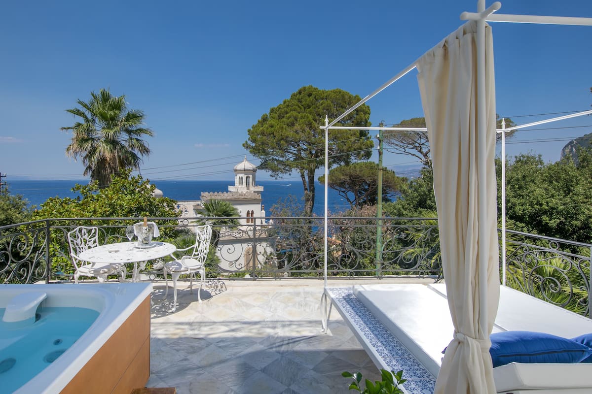 Un balcón con vista al mar, una mesa blanca con sillas, una piscina pequeña y una pérgola.