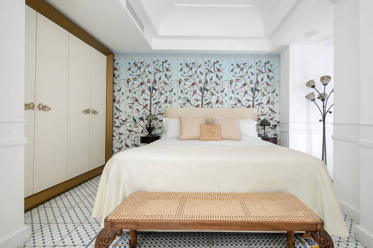 Un dormitorio con una cama grande, un banco, un papel tapiz estampado y una planta alta.
