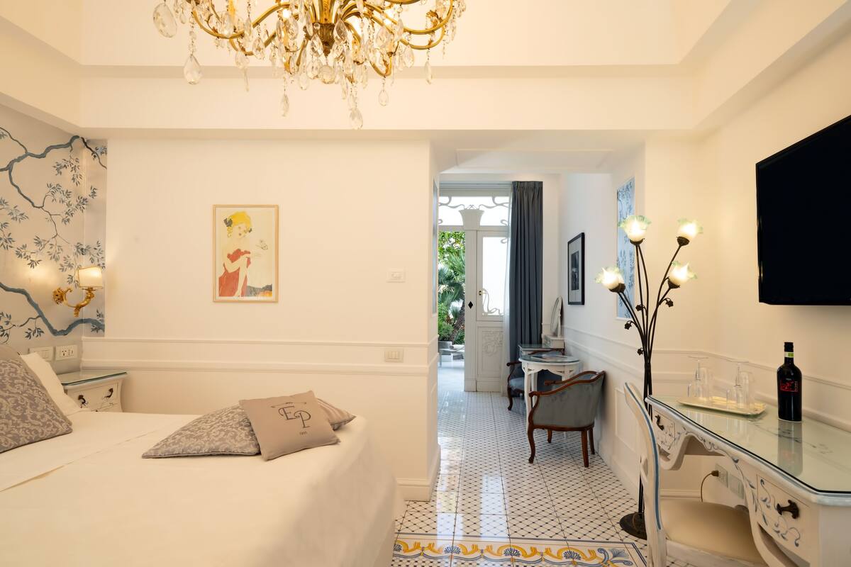 A hotel room with a bed, a desk, a chair, a chandelier, and a TV on the wall.