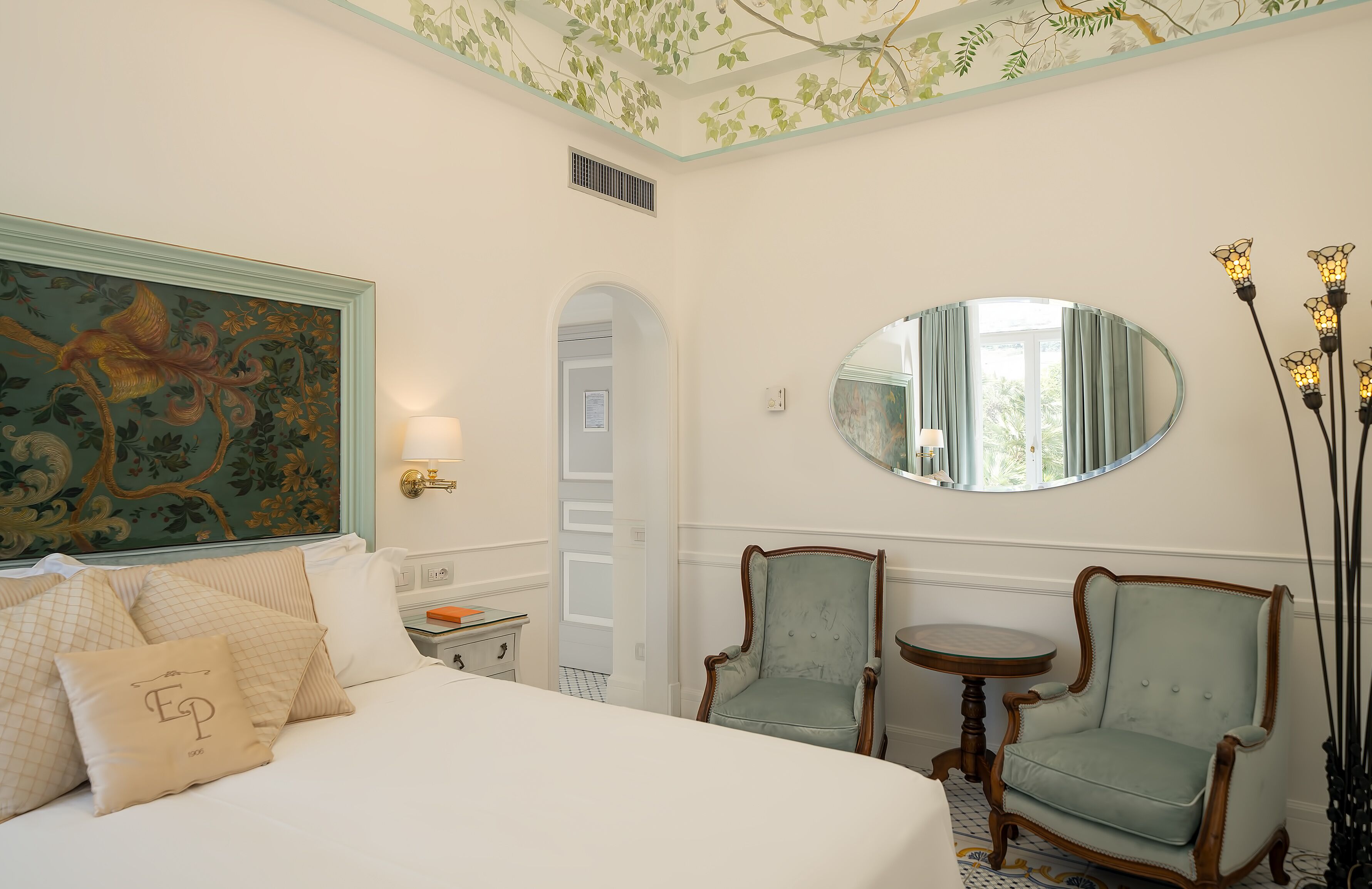 A hotel room with a large bed, two armchairs, a round table, a mirror, and a ceiling mural.