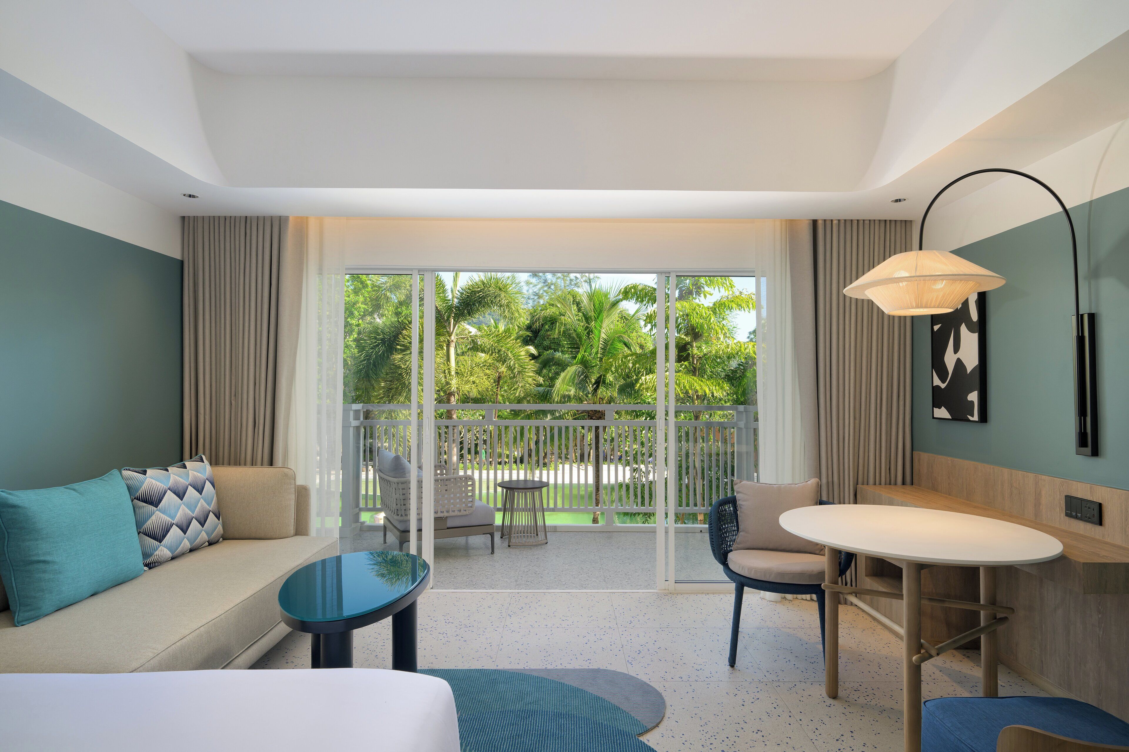 Ein Hotelzimmer mit Sofa, kleinem Tisch, Stuhl und Balkon mit Blick auf Grünflächen.