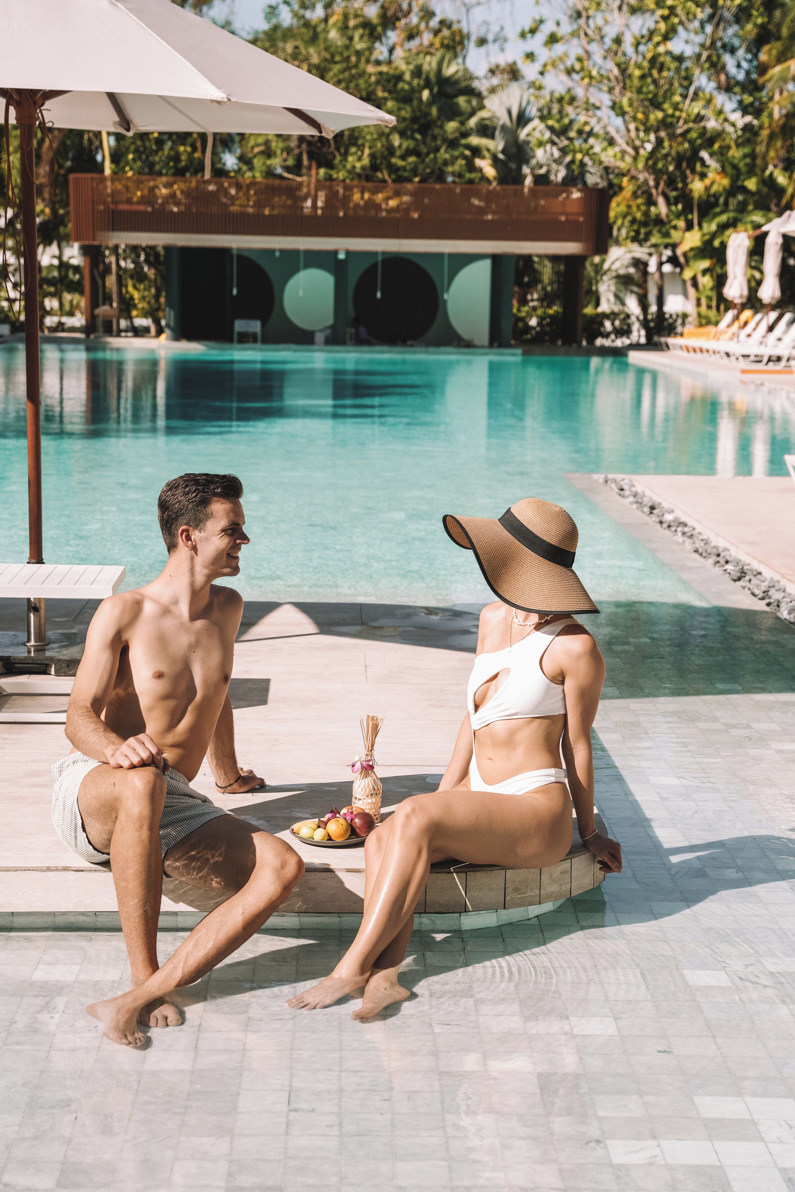 Zona de piscina con una tumbona y una sombrilla, una mesita con fruta y una piscina al fondo.
