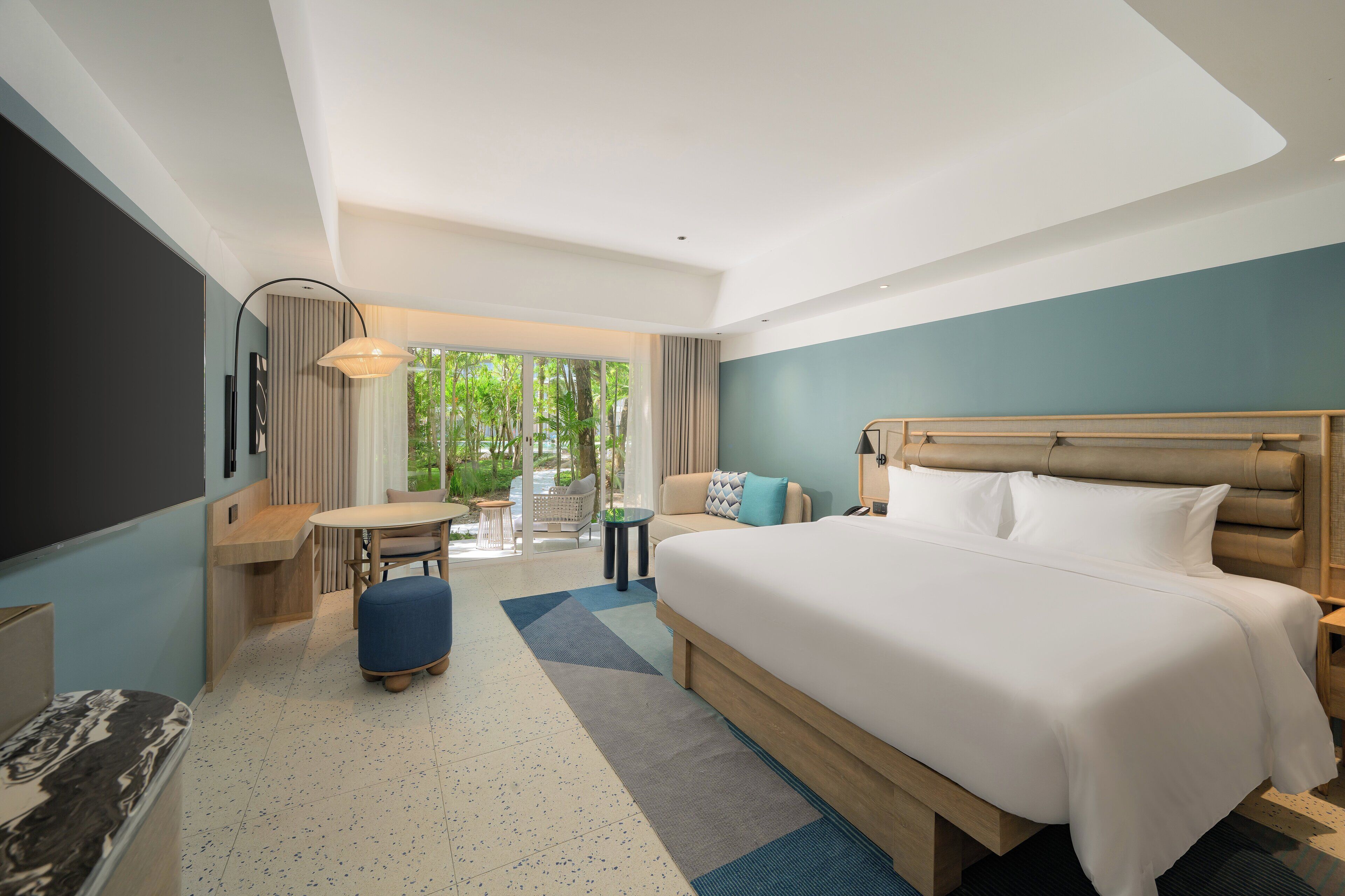 A modern hotel room with a large bed, a desk, and a view of greenery.