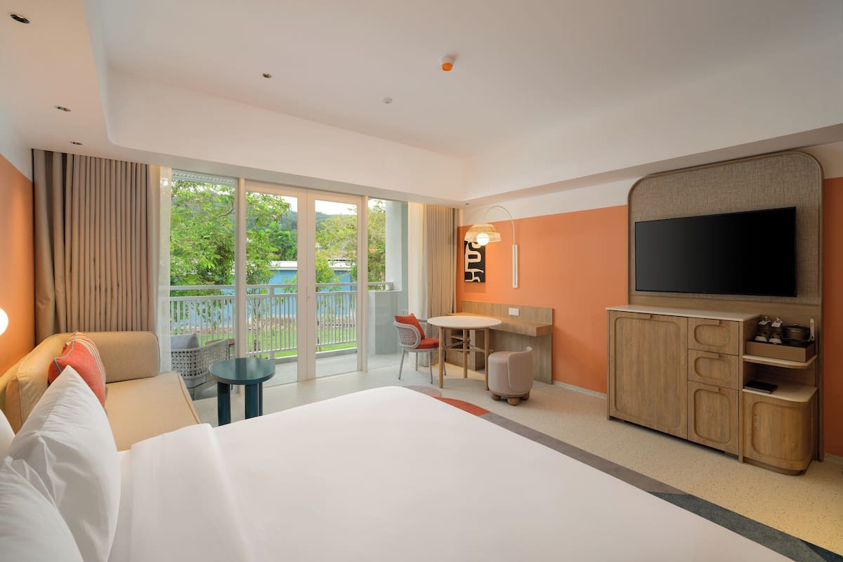 A hotel room with a large bed, a desk with a chair, a television, and a balcony with a view of greenery.