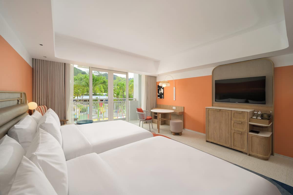 A hotel room with two beds, a TV, a desk, and a view of palm trees.
