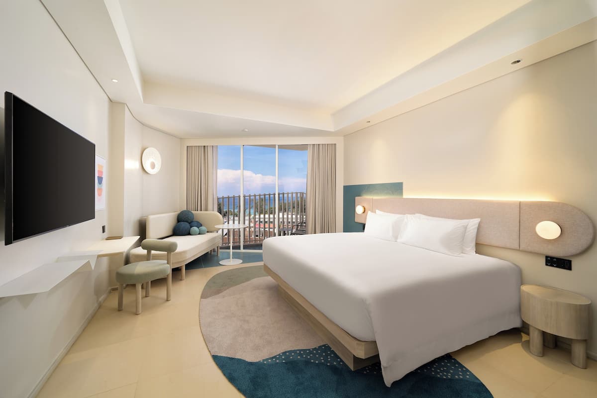 A modern hotel room with a large bed, a flat-screen TV, a sofa, and a balcony with a view.