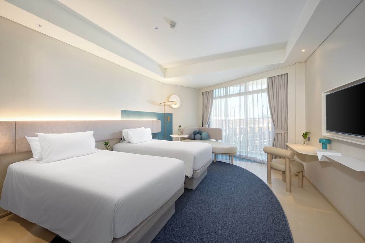 A hotel room with two beds, a desk, a chair, a TV, and a large window.
