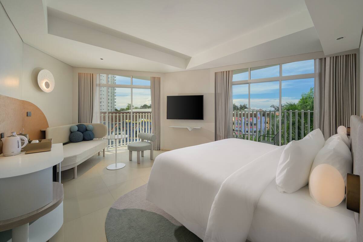 A modern hotel room with a large bed, a desk with a chair, a TV, and a balcony with a view.