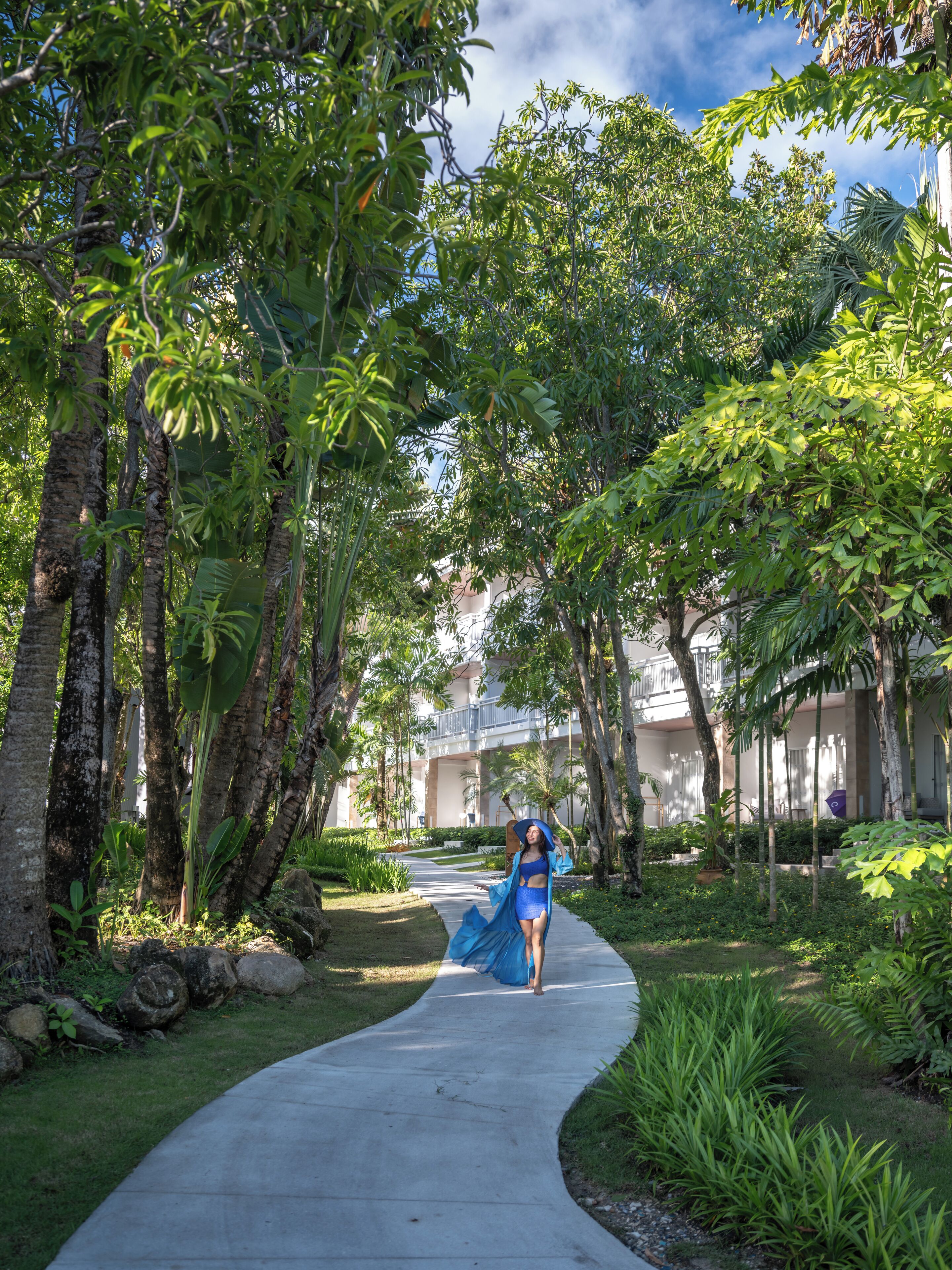 A pathway lined with tropical trees leading to a building with balconies.