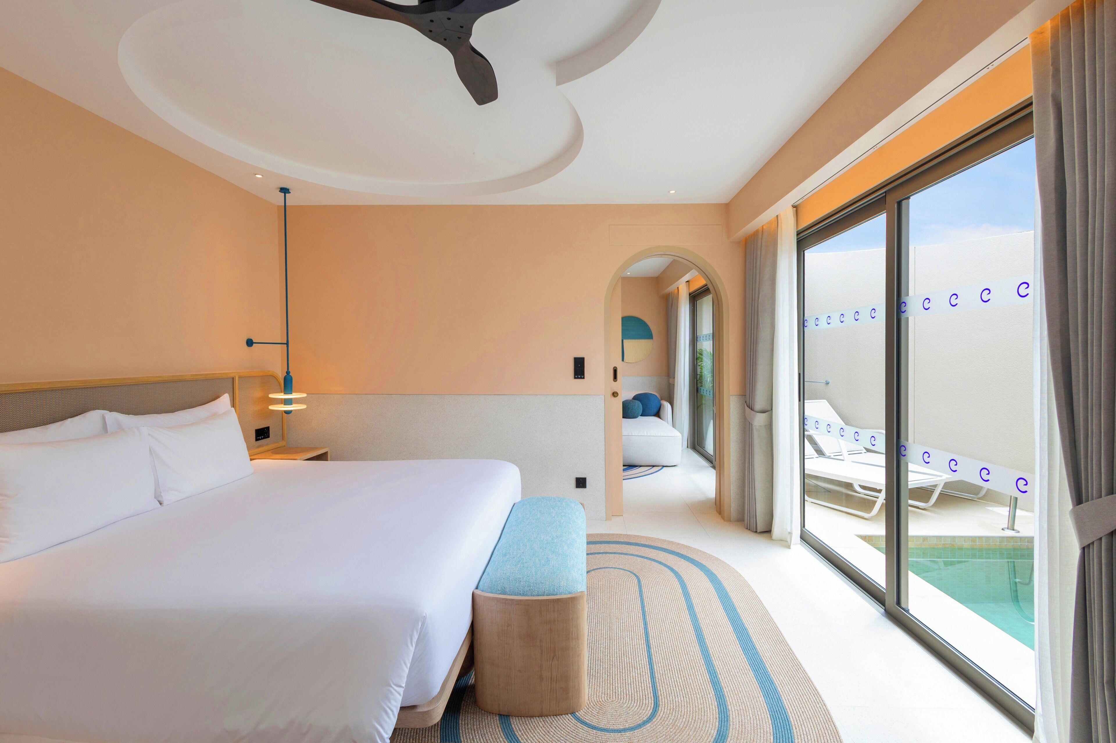 A hotel room with a large bed, a bench, a ceiling fan, and a view of a pool area.