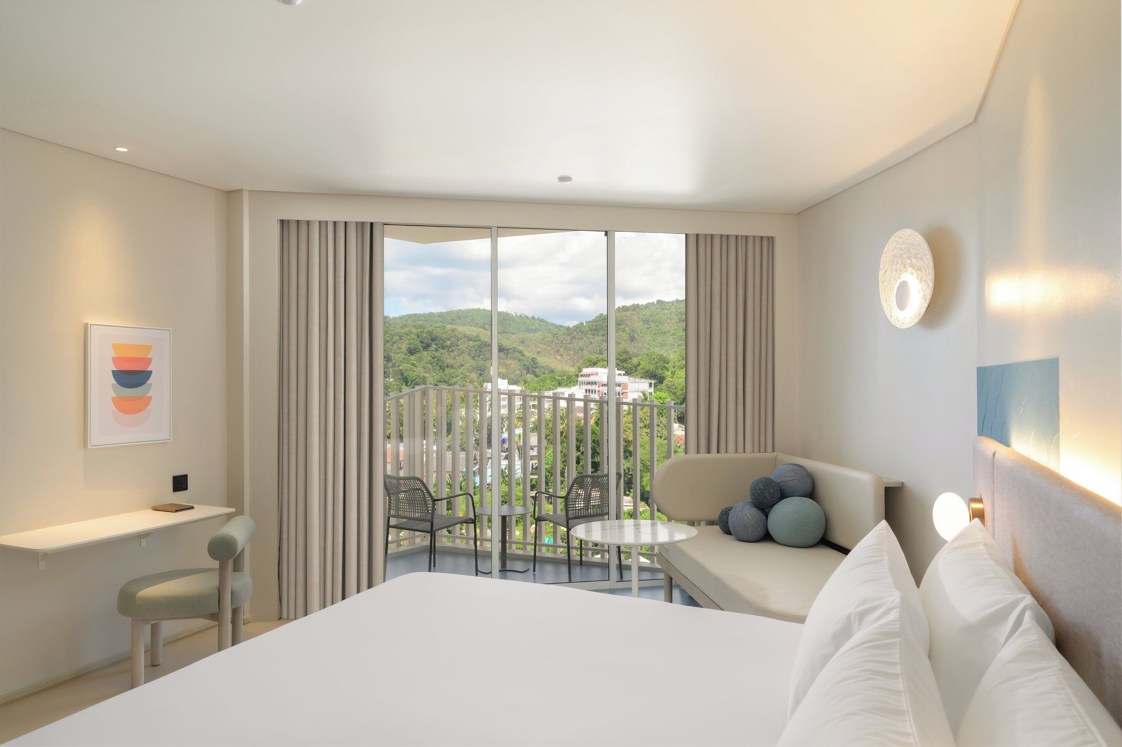 Hotelkamer met een groot bed, een bureau, een stoel, een balkon met uitzicht op de heuvels en een wandlamp.