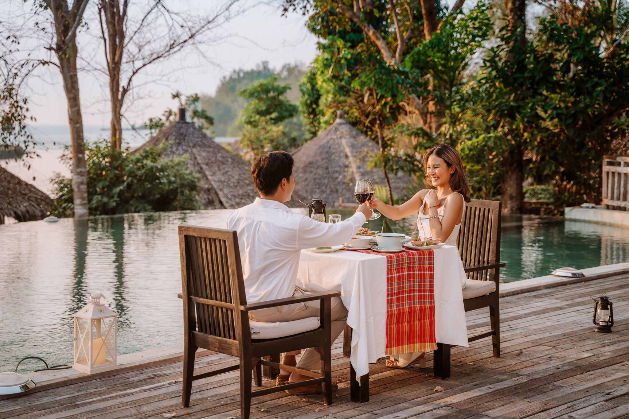 Una pareja cenando junto a una piscina con vista al mar y cabañas de techo de paja.