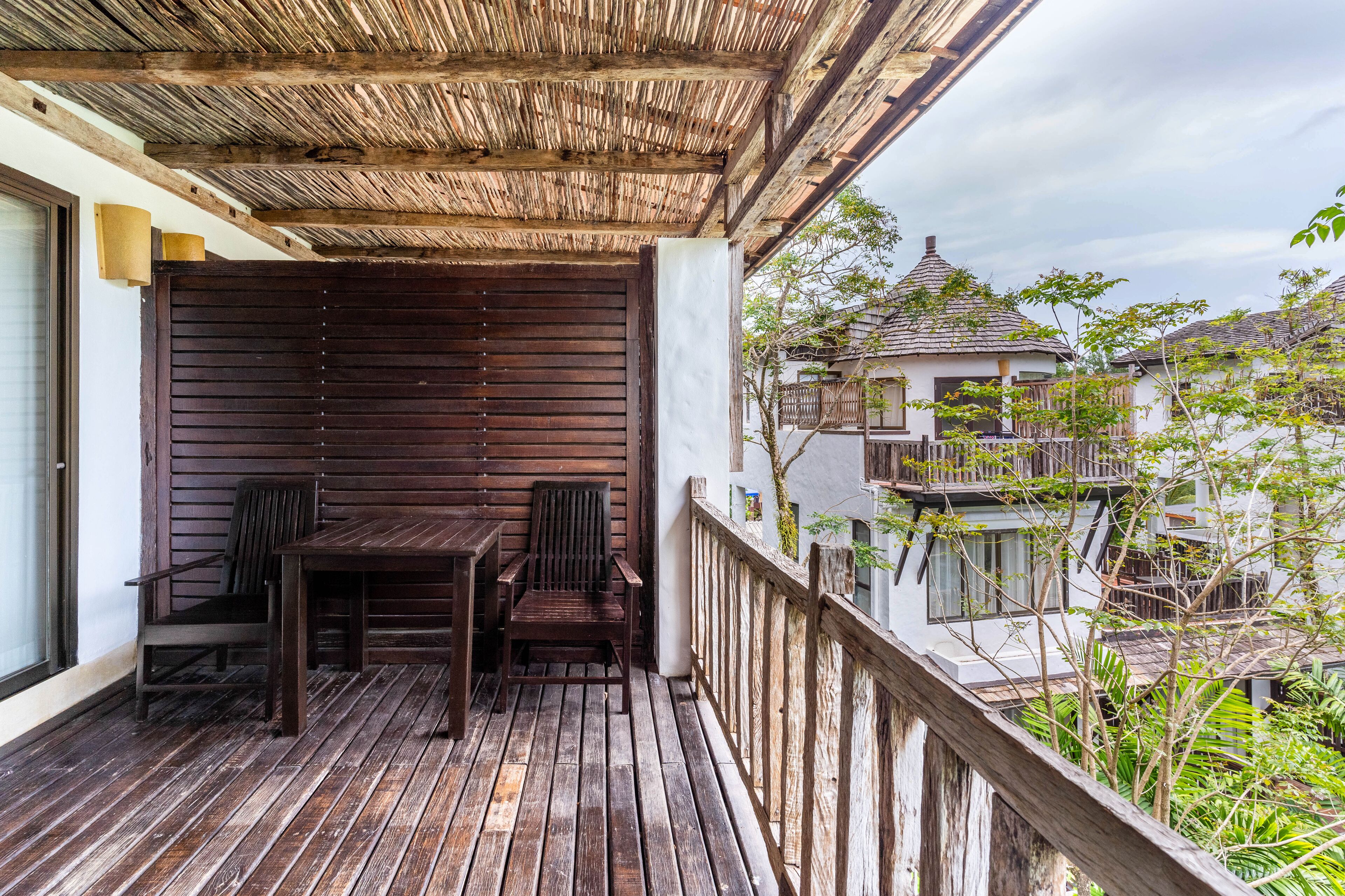 Een houten terras met een tafel en stoelen, een rieten dak en uitzicht op naburige gebouwen.