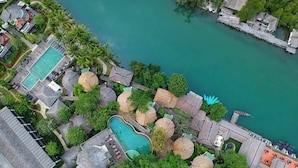An aerial view of a resort with a swimming pool, thatched-roof structures, and a dock extending into a turquoise body of water. - Aana Resort & Spa (Ko Chang)