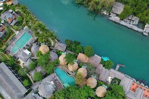 Vista aérea de un resort con piscina, estructuras de techo de paja y un muelle que se adentra en una masa de agua turquesa.