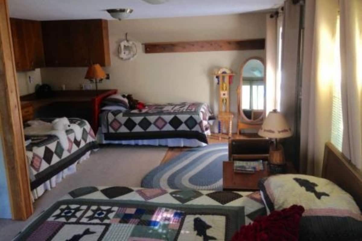 Une chambre de taille modeste, équipée de deux lits, d’une petite kitchenette, d’un miroir et d’une fenêtre avec des rideaux.