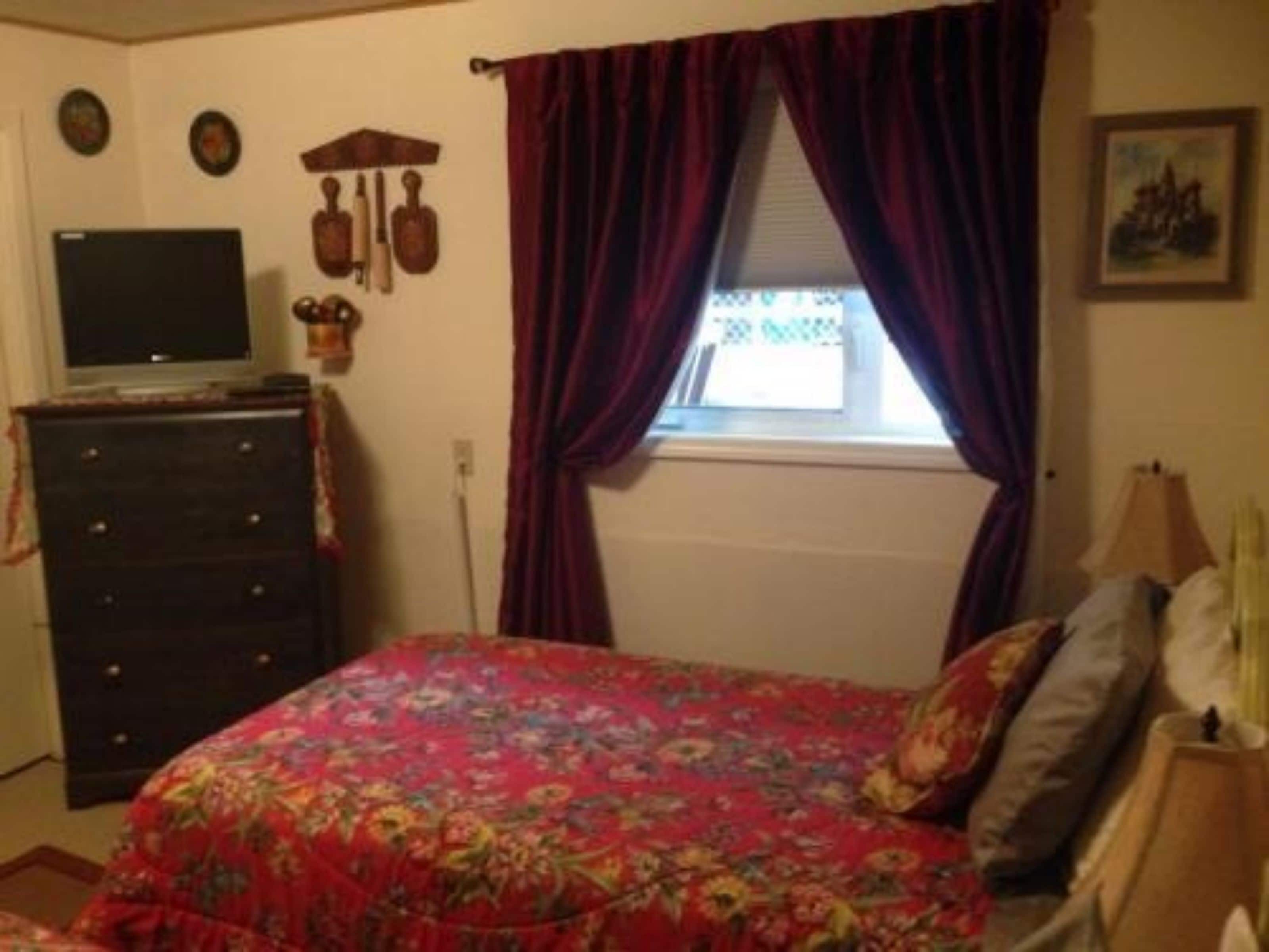 A bedroom with a floral bedspread, a dresser, a television, and a window with curtains.