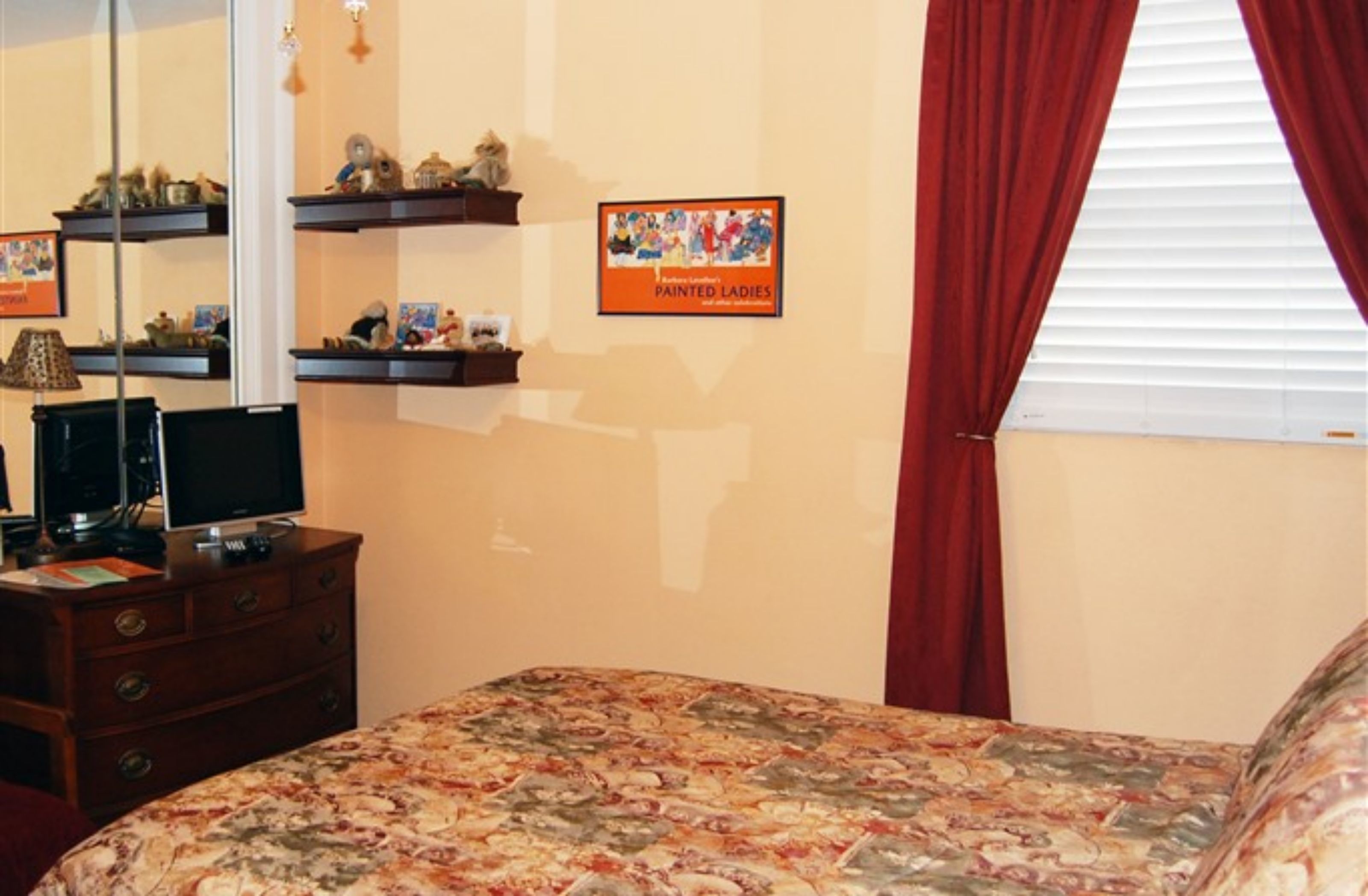 A bedroom with a bed, a dresser, a TV, shelves with decorative items, and a window with red curtains.