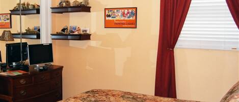 A bedroom with a bed, a dresser, a TV, shelves with decorative items, and a window with red curtains.