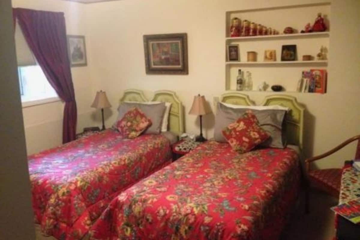 Une chambre avec deux lits simples, chacun agrémenté de draps à motifs floraux et de lampes de chevet. On y trouve une étagère avec des objets décoratifs et un tableau encadré au mur.