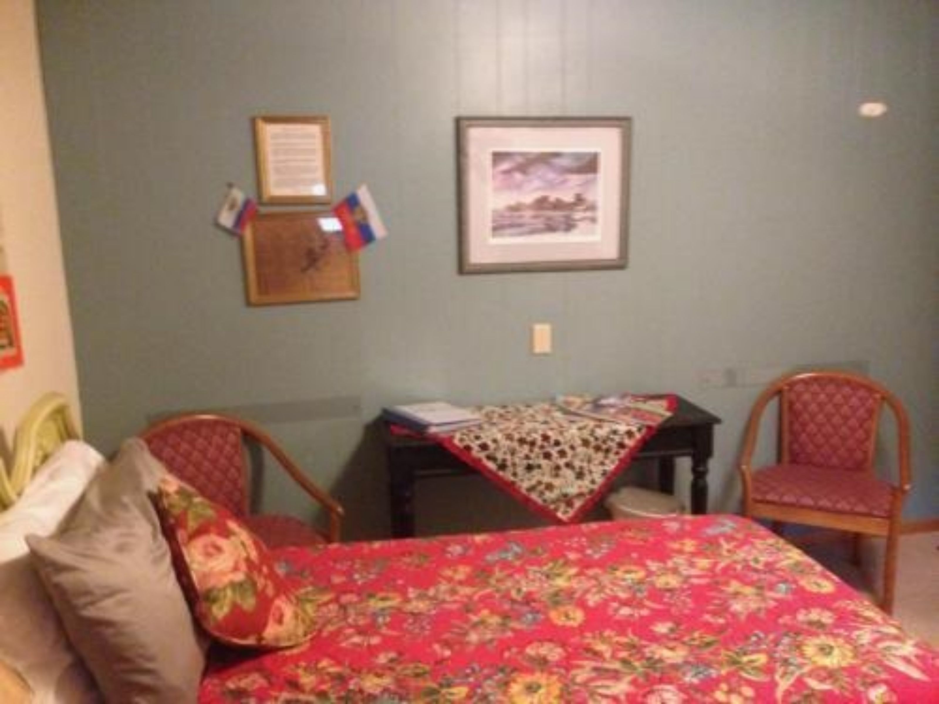 Ein Zimmer mit einem Bett, zwei Stühlen, einem Tisch und einer Wand mit gerahmten Gegenständen.