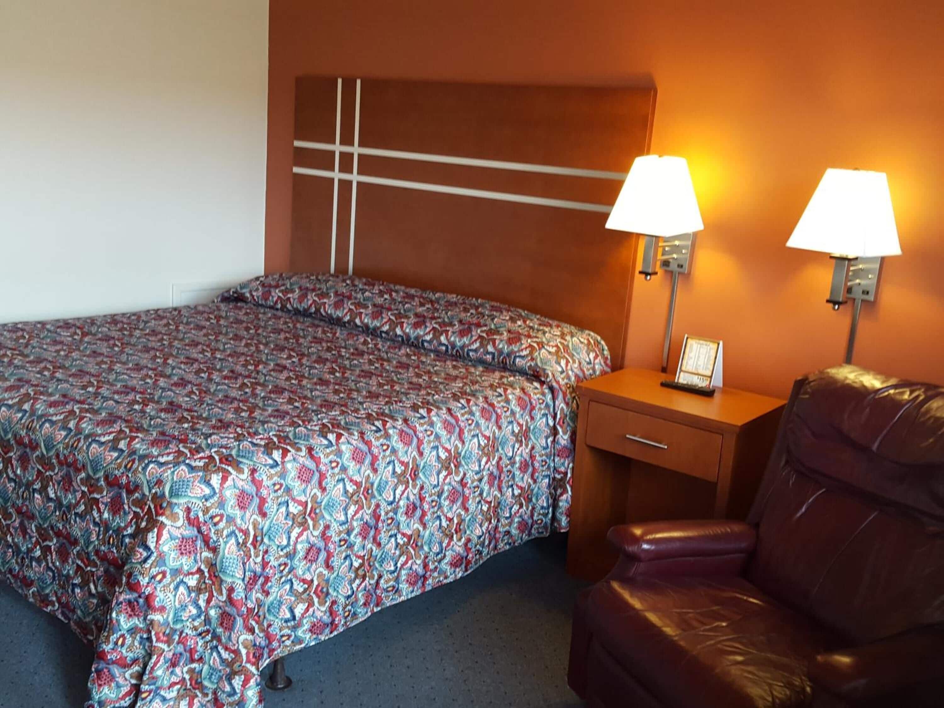 Une chambre d’hôtel comprenant un lit, deux lampes de chevet, une table de chevet, un fauteuil en cuir et une horloge murale.