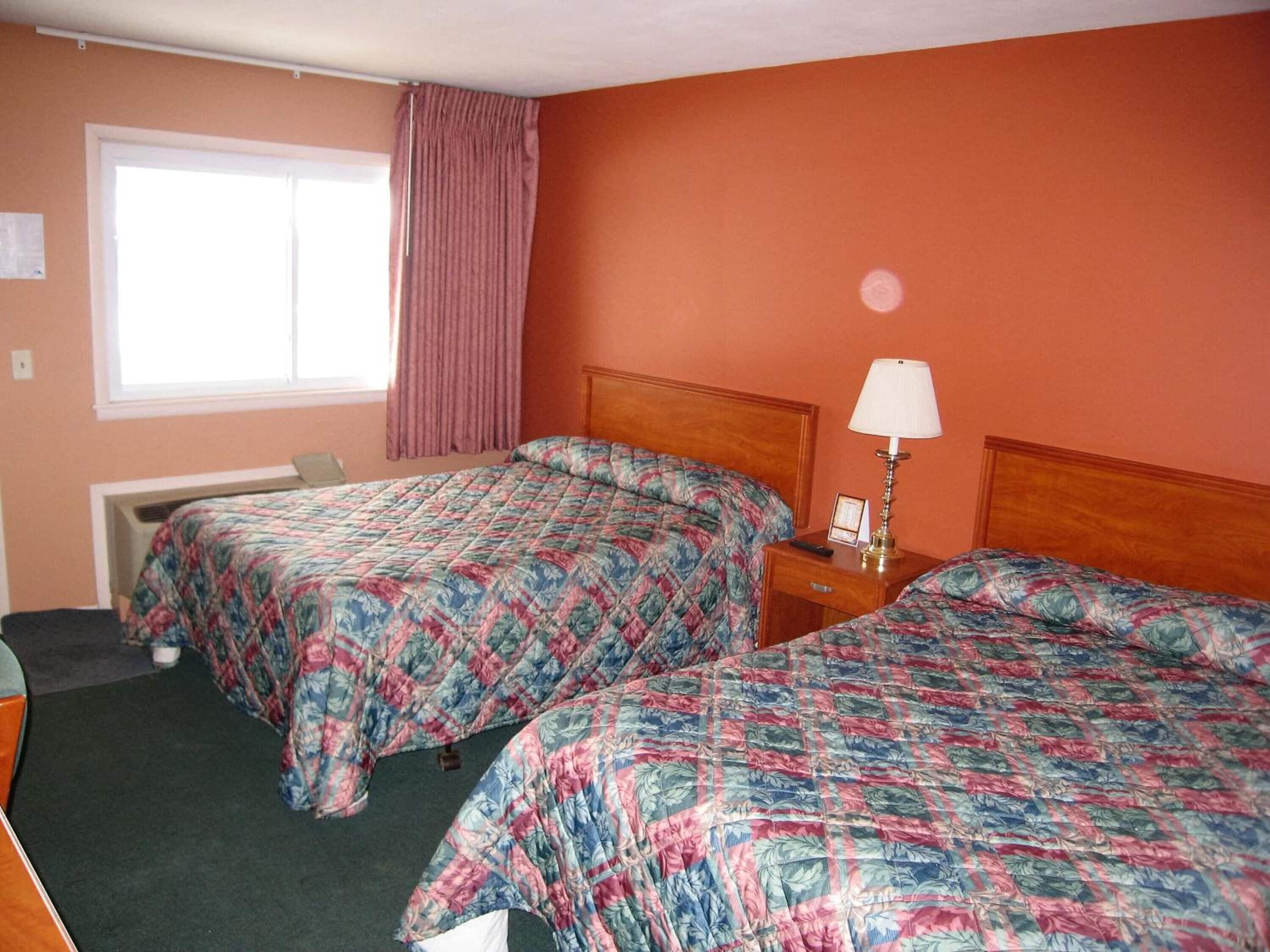 Quarto de hotel com duas camas, uma janela com cortinas, uma mesa de cabeceira com abajur e um radiador.