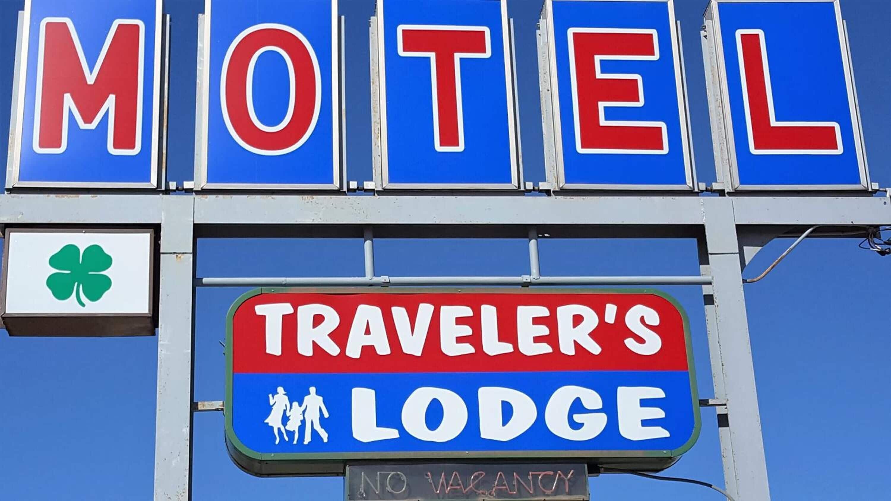 Letrero de motel con el texto 'MOTEL' y 'TRAVELER'S LODGE', un símbolo de trébol de cuatro hojas y un letrero de 'NO VACANCY'.