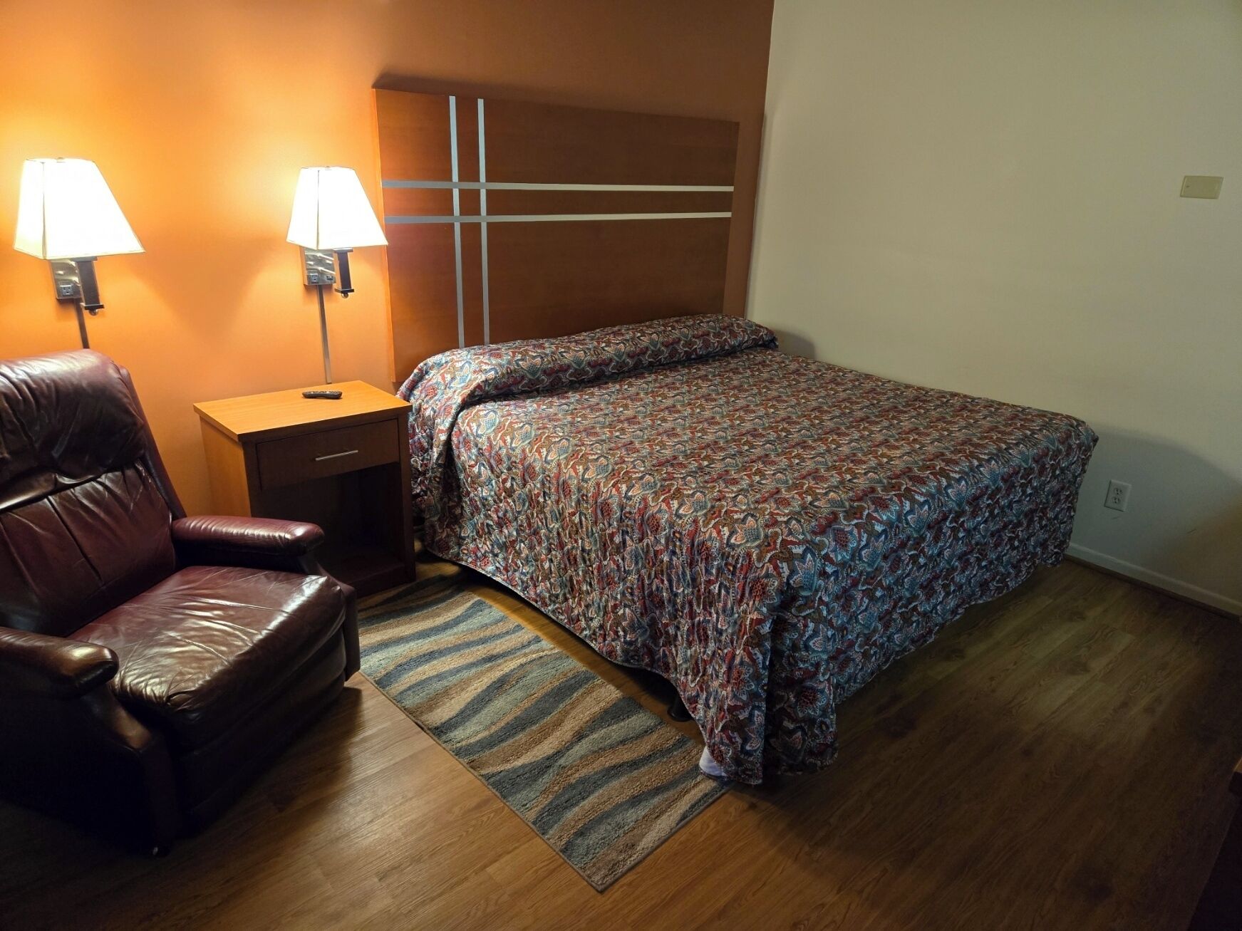 A bedroom with a bed, a leather chair, a nightstand, two wall lamps, and a patterned rug.
