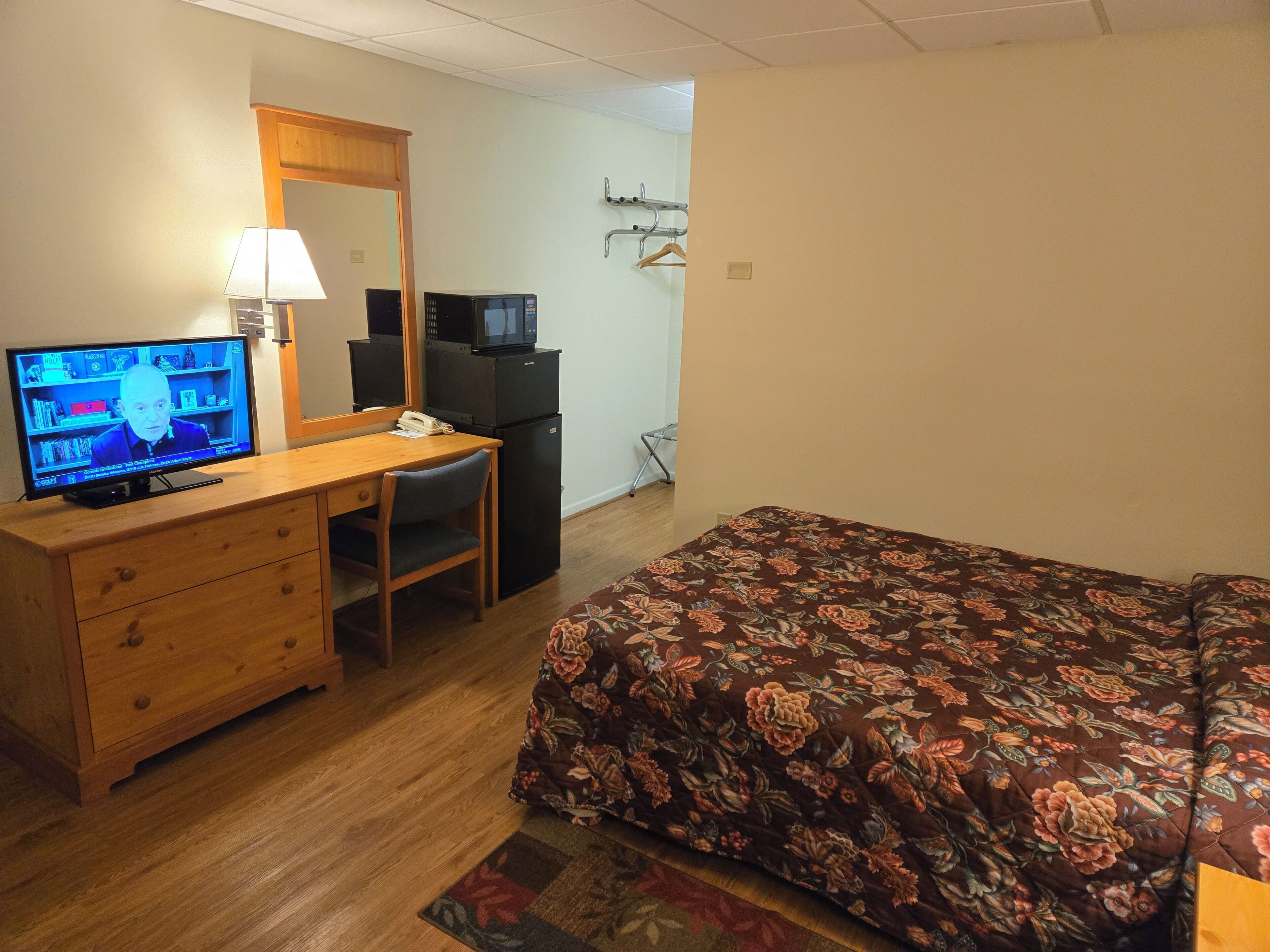A hotel room with a wooden desk, a television, a bed with a floral bedspread, and a small kitchenette.