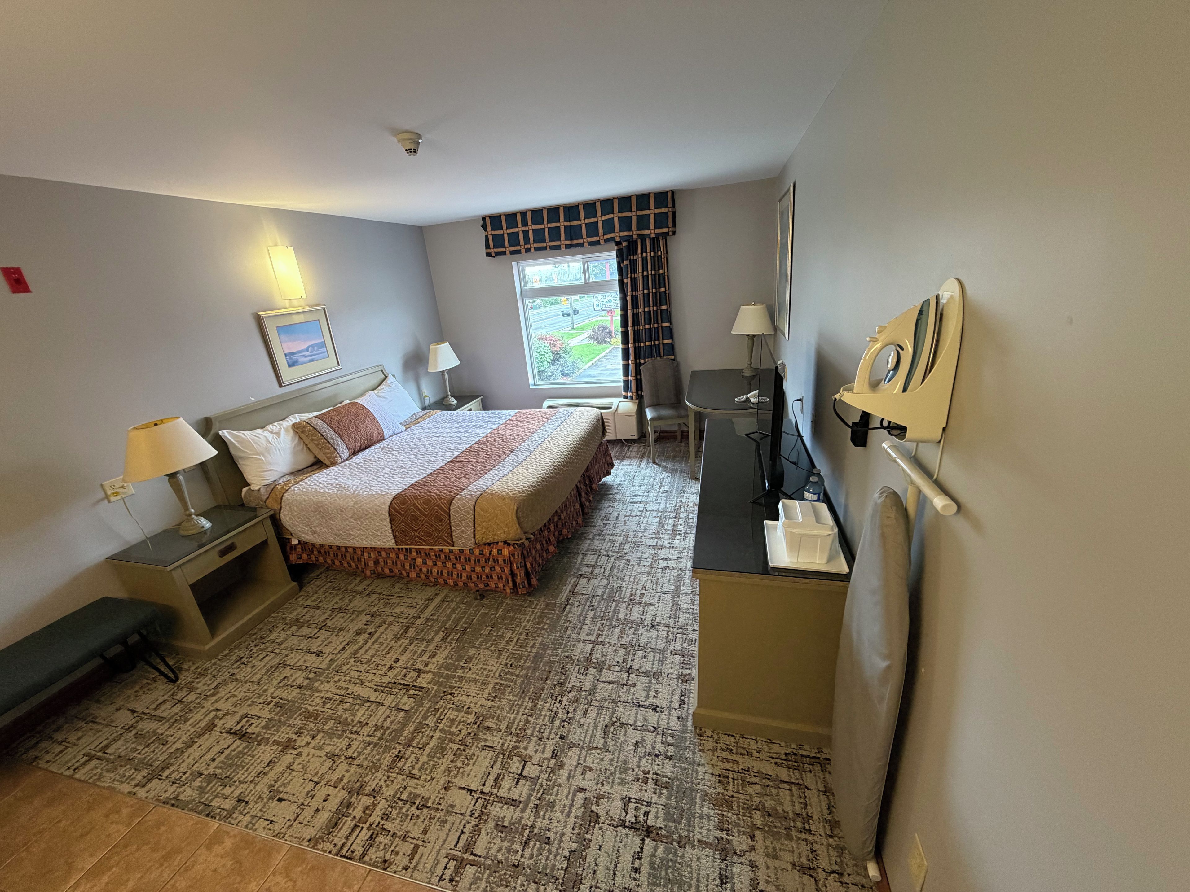 A hotel room with a bed, bedside tables, a desk, a chair, a lamp, a radiator, and a window with curtains.