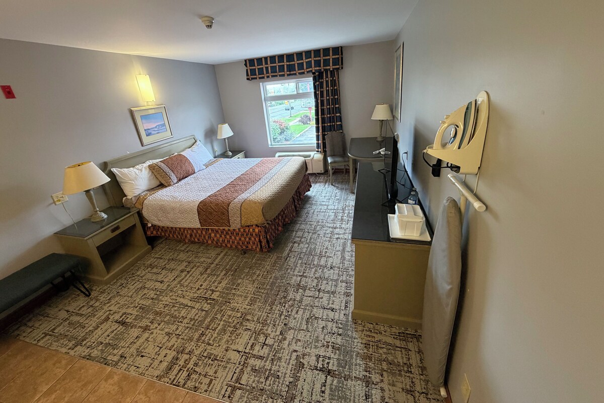 A hotel room with a bed, bedside tables, a desk, a chair, a lamp, a radiator, and a window with curtains.