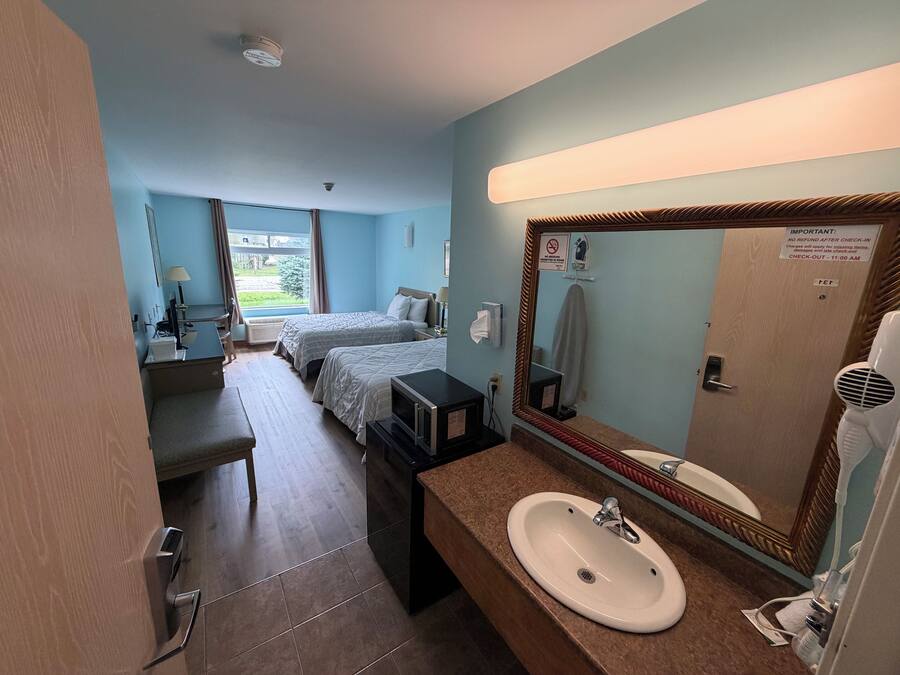 A hotel room with two beds, a bathroom with a sink and mirror, and a window with curtains.