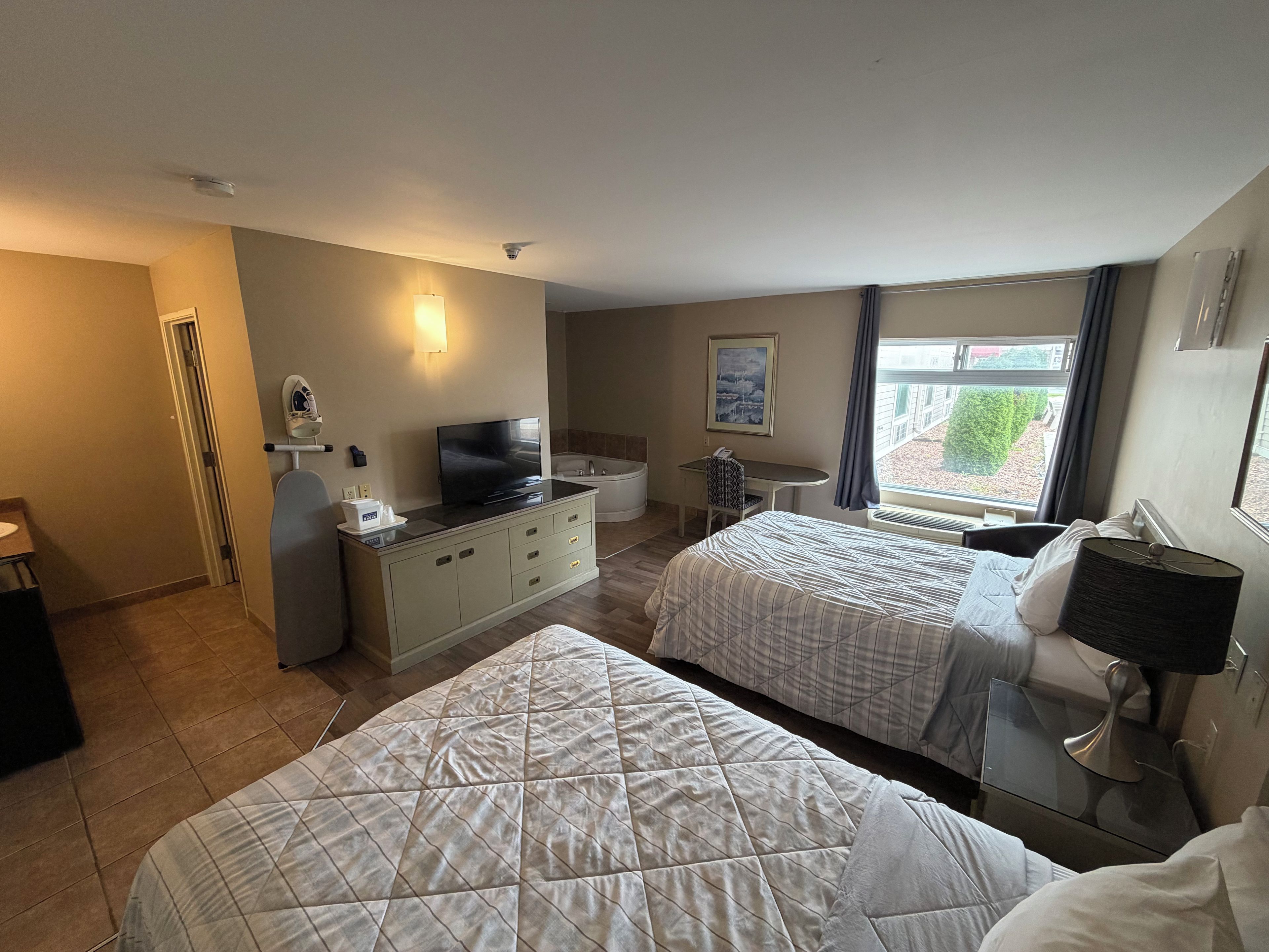 A hotel room with two beds, a desk with a flat-screen TV, a small bathroom, and a window with a view.
