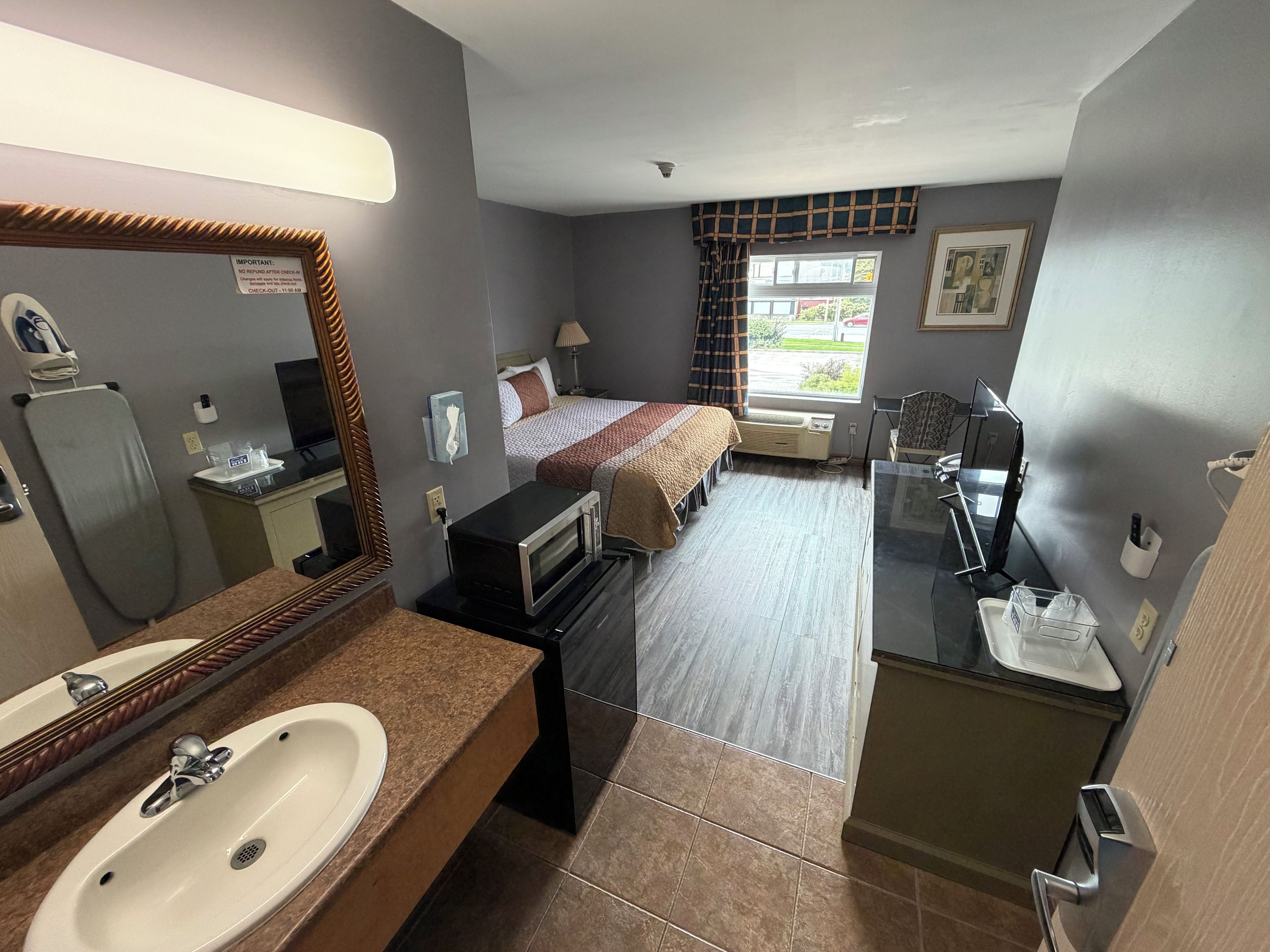 A hotel room with a bed, a bathroom with a sink and mirror, a microwave, and a window with curtains.