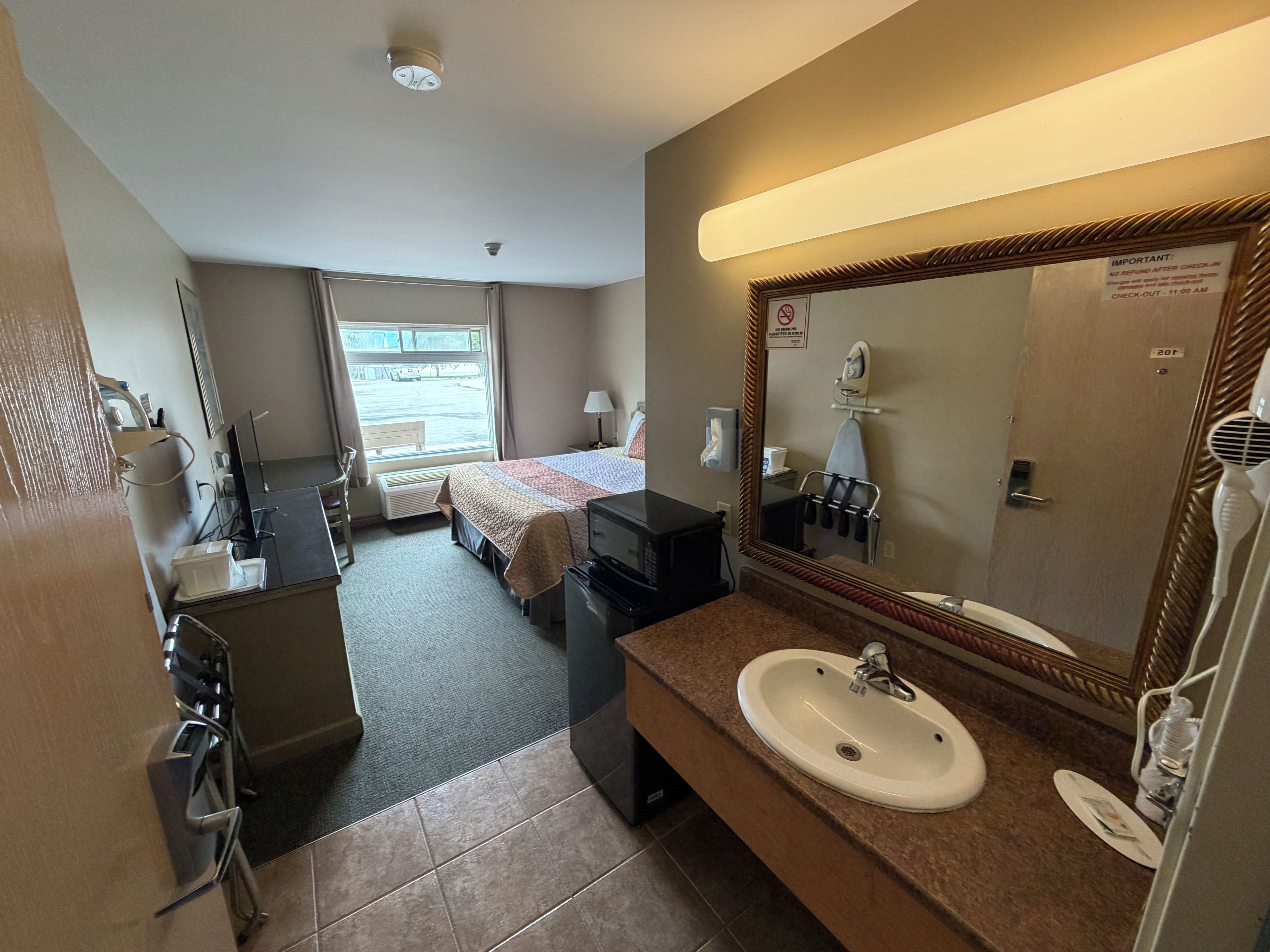 Habitación de hotel con cama, escritorio, cómoda, baño con lavamanos y espejo, y ventana con vistas al exterior.