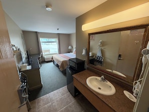 Ett hotellrum med en säng, ett skrivbord, en byrå, ett badrum med handfat och spegel samt ett fönster med utsikt över omgivningen.