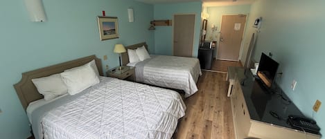 A hotel room with two beds, a desk with a computer, a TV, and a small kitchenette.