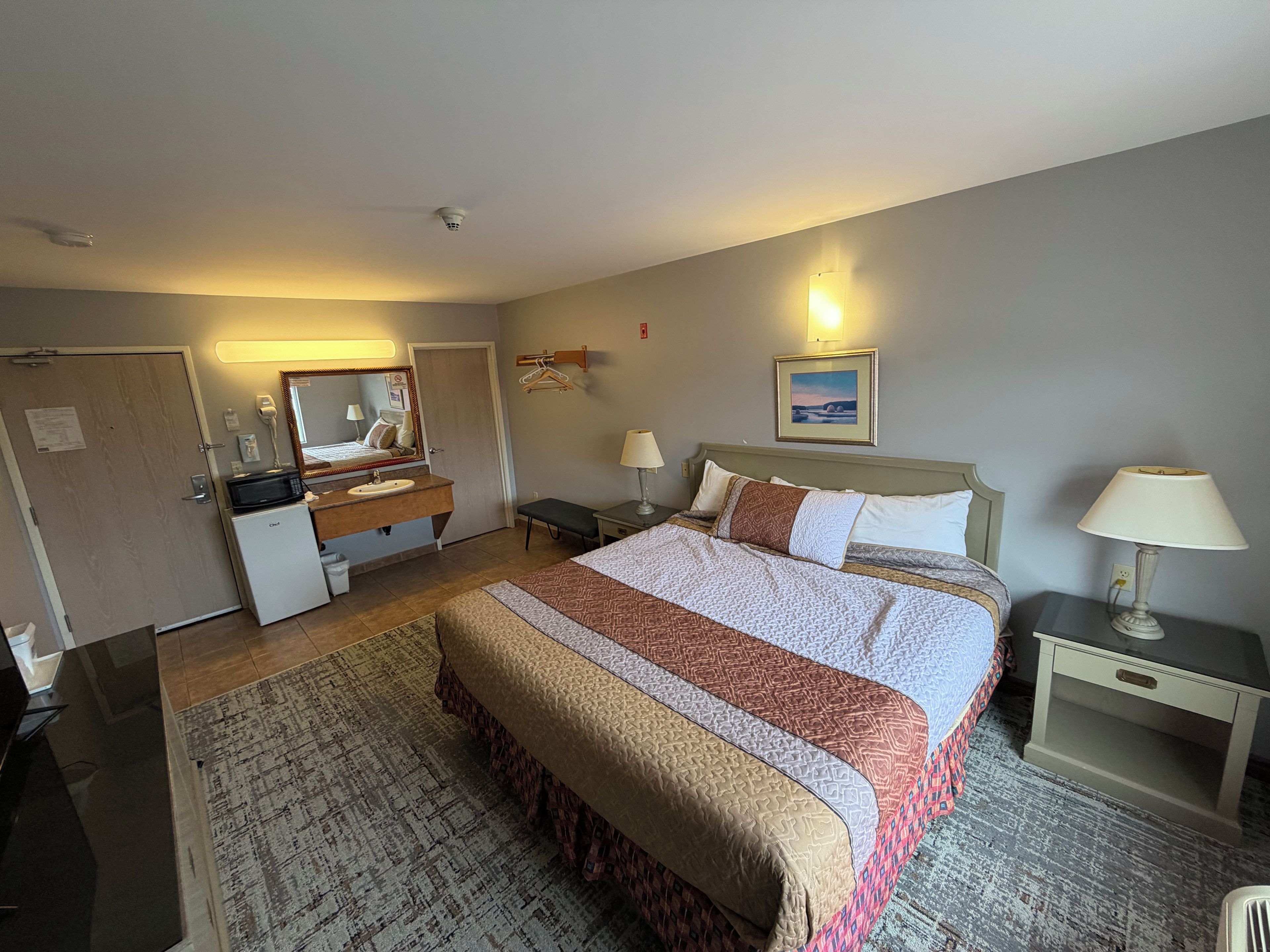 A hotel room with a bed, bedside tables, a desk, a mirror, a small refrigerator, and a wall-mounted lamp.