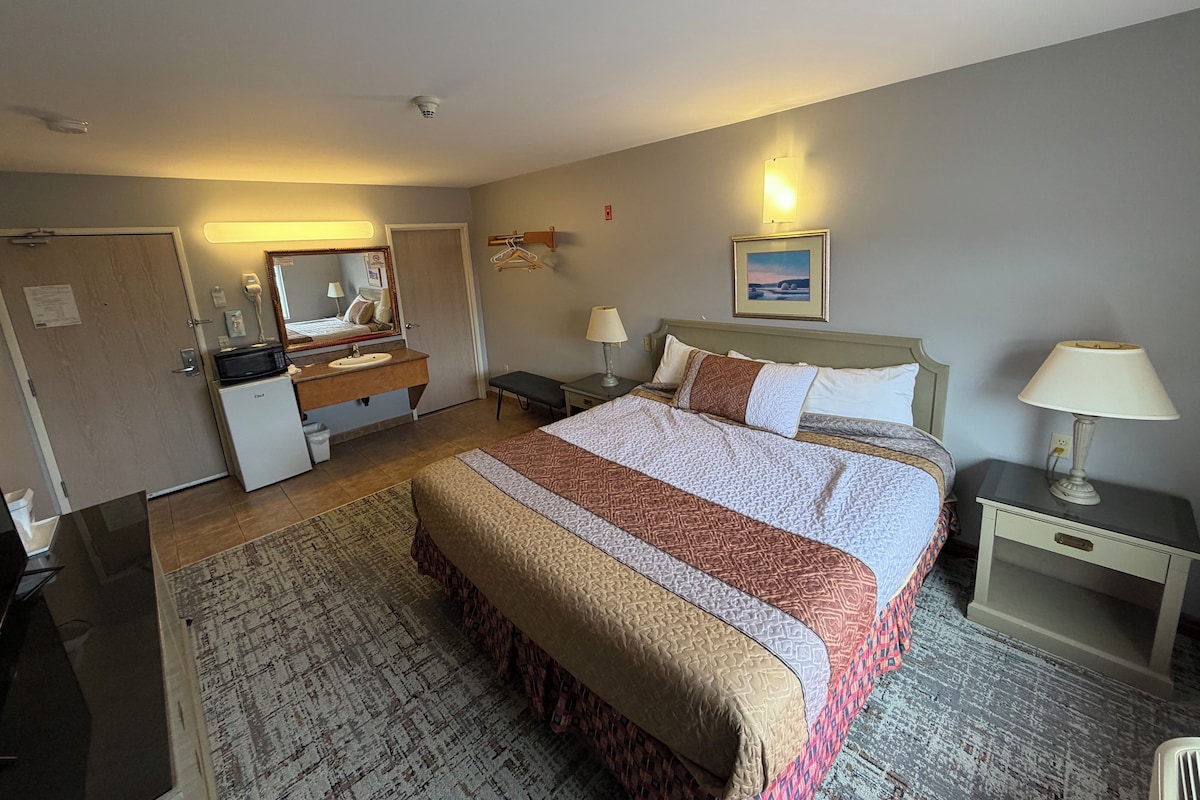 A hotel room with a bed, bedside tables, a desk, a mirror, a small refrigerator, and a wall-mounted lamp.