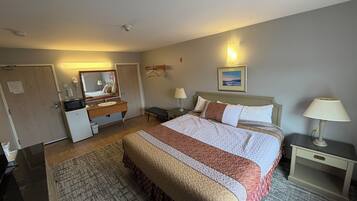 A hotel room with a bed, bedside tables, a desk, a mirror, a small refrigerator, and a wall-mounted lamp.
