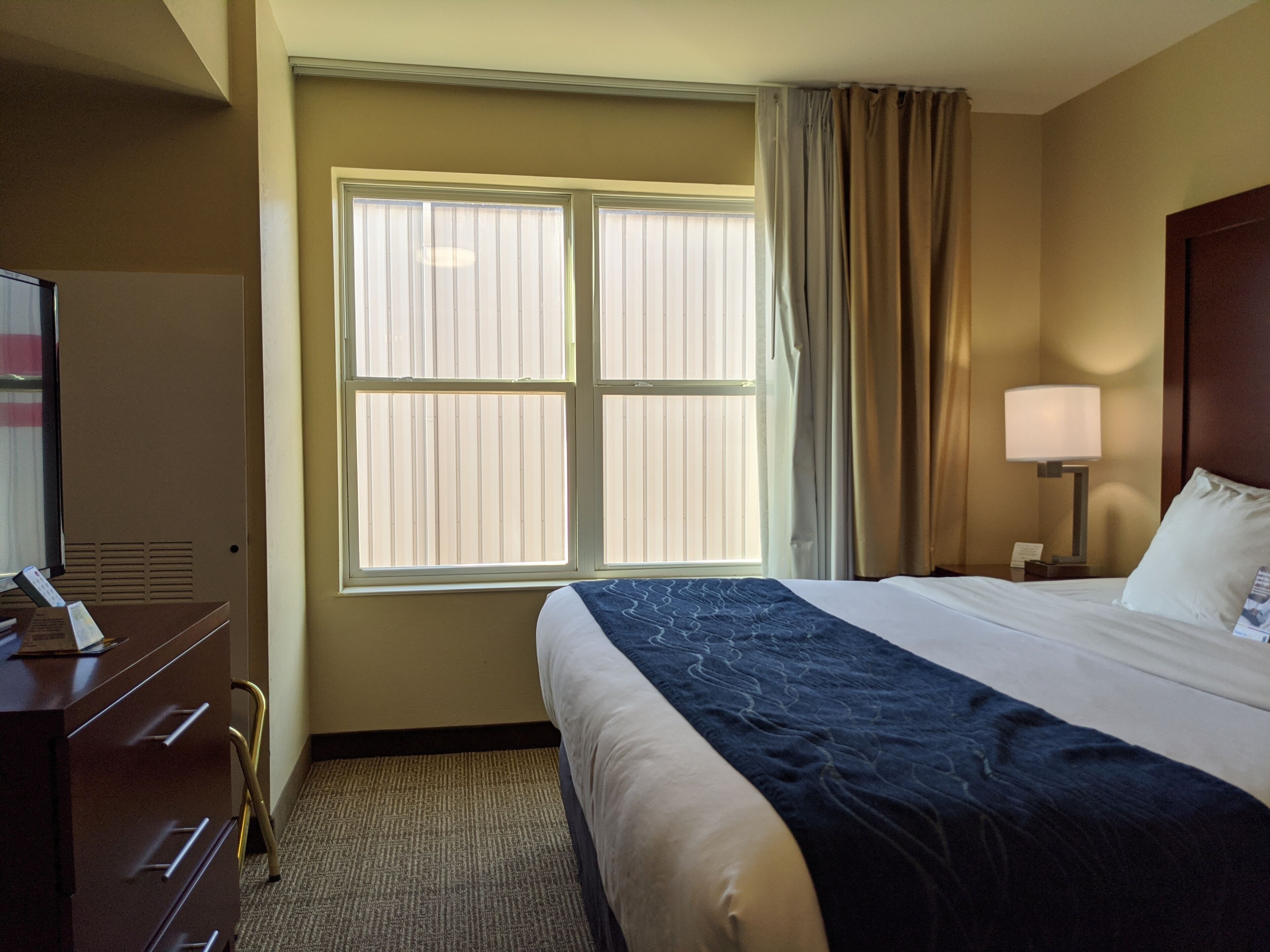 A hotel room with a large bed, a desk with a lamp, a window with blinds, and a dresser.