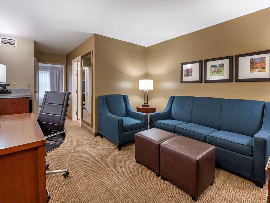 A hotel room with a desk, two blue armchairs, a sofa, and a brown ottoman. There are three framed pictures on the wall.