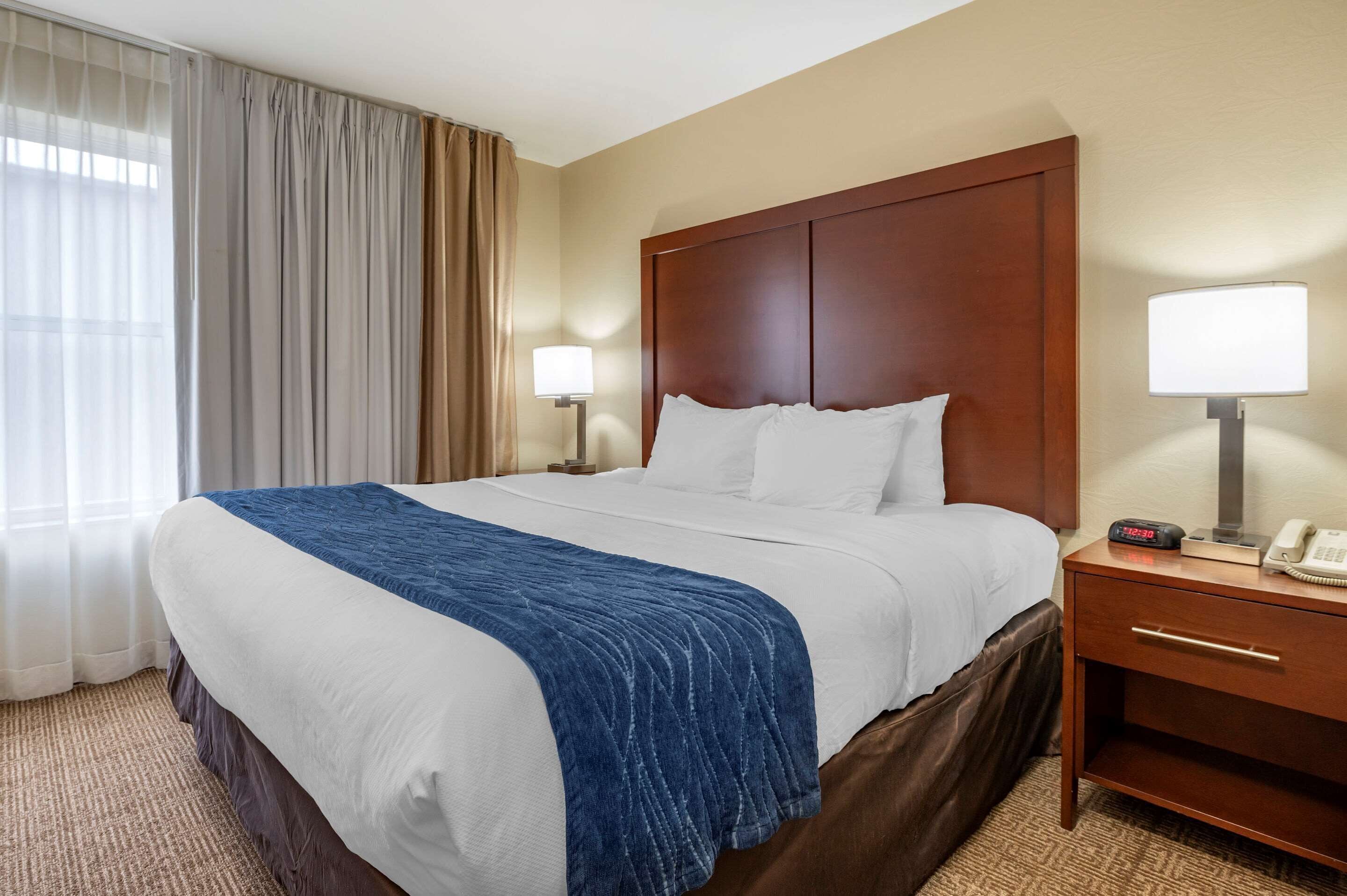 Quarto de hotel com uma cama grande, cabeceira de madeira, duas mesas de cabeceira com luminárias, relógio na cabeceira e piso coberto por carpete.