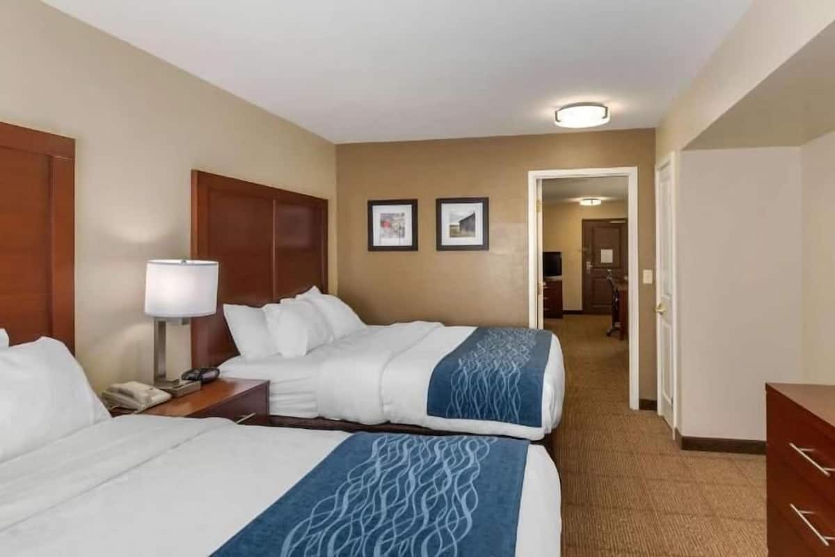 A hotel room with two beds, a TV, a desk, a lamp, and a door leading to another room.